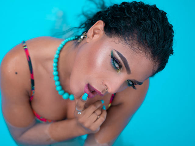 VioletHotX met krullend zwart haar, groene oogmake-up, een turquoise kralenketting en een kleurrijke bikini-top, met turquoise lippenstift op, een neusring, op een blauwe achtergrond, kijkt omhoog met een sexy uitdrukking..