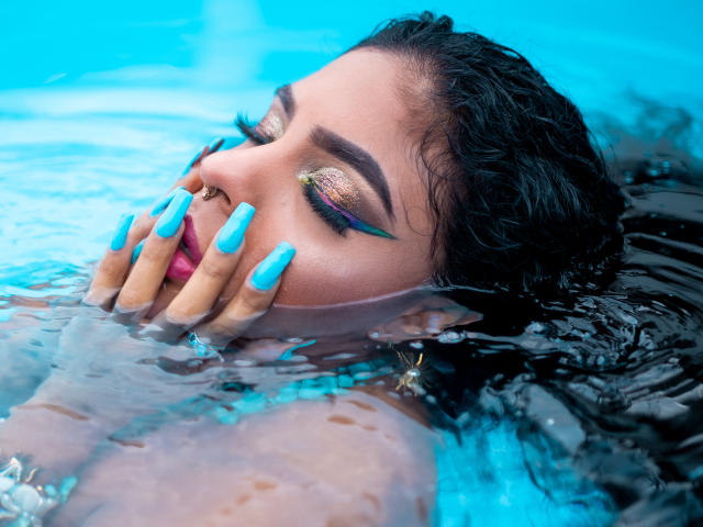 VioletHotX's face, half-submerged in blue water, with black hair, gold eyeshadow, blue nails, nose ring, and pink lips, hands covering her mouth. Her eyes are closed.
