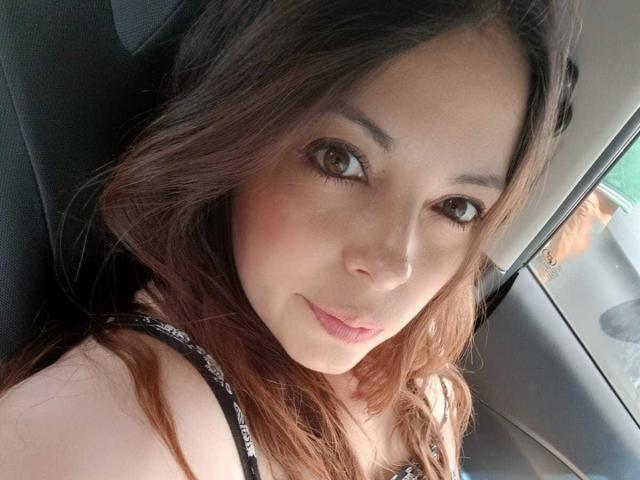 Seher has brown eyes, light skin, and wavy brown hair. She's wearing a black strappy top, looking at the camera with a slight smile. Seher's tits are partially visible.
