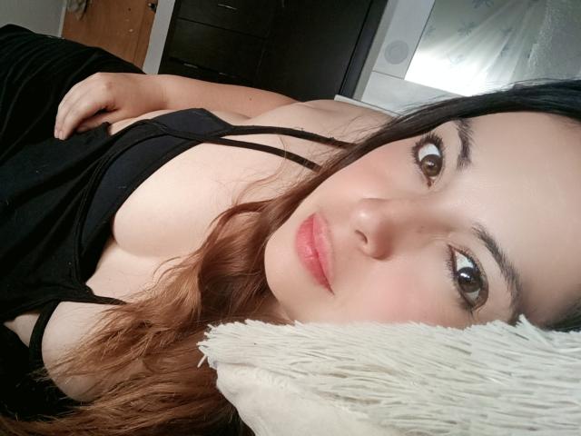 Seher with wavy brown hair, black tank top, lying on a white pillow, showing her boobs, pink lips, brown eyes, and soft expression in a relaxed, intimate shot.