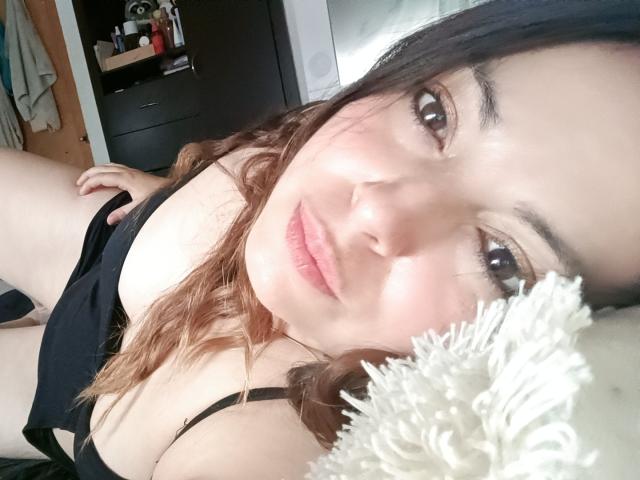 Seher, avec ses cheveux bruns ondulés, sa peau claire et ses yeux marrons, est allongée en lingerie noire, montrant sa poitrine, avec un objet blanc et duveteux au premier plan..