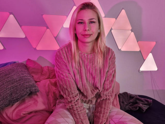 ViviVanilli-hot with blonde hair, pink sweater, and white pants, sitting on a bed, smiling, pink and purple lighting, geometric wall decorations in the background.