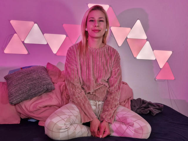 ViviVanilli-hot with pink-tinted hair, wearing a sheer pink long-sleeve top and white plaid pants, sitting on a bed with pink and gray pillows. Pink triangular lights behind.