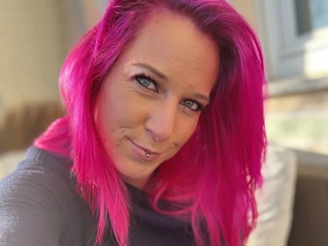 Candy-bitch-hot with bright pink hair, gray eyes, and a nose piercing, smiling at the camera. She's wearing a gray turtleneck, and her tits are subtly visible.