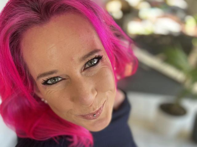 Candy-bitch-hot with bright pink hair, green eyes, and a subtle smile, looking up with a small lip piercing, wearing a dark top, in a blurry background.