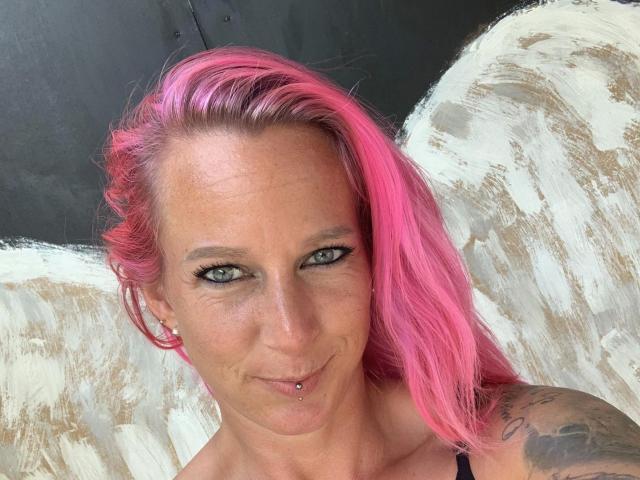 Candy-bitch-hot with pink hair, green eyes, pierced lip, and tattooed arm, smiling at the camera, wearing a black strap. Boobs visible, small earrings, and a confident look.