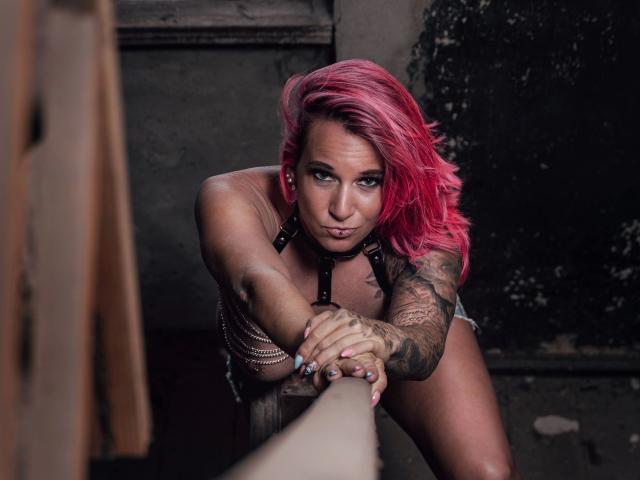 Candy-bitch-hot with pink hair, tattoos on her arm, leaning on a railing, wearing a black leather top, pouting with intense eyes, showing her tits and a sexy, confident look.