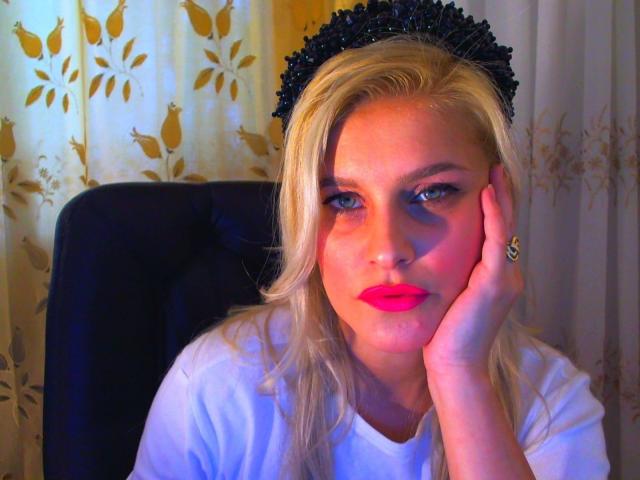KillerSmilexxx-hot with blonde hair, black spiked headpiece, pink lipstick, white shirt, hand on cheek, ring on finger, blue and orange lighting, sitting in black chair.