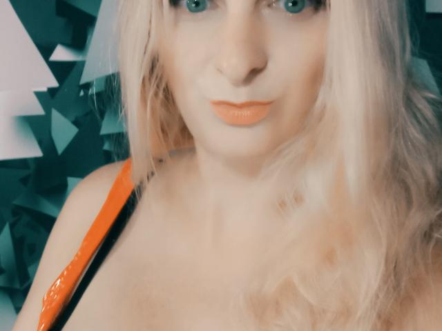 MonicaMiss has blonde hair, blue eyes, orange lipstick, and is wearing an orange strap. Her tits are partially visible. She looks straight at the camera with a neutral expression.