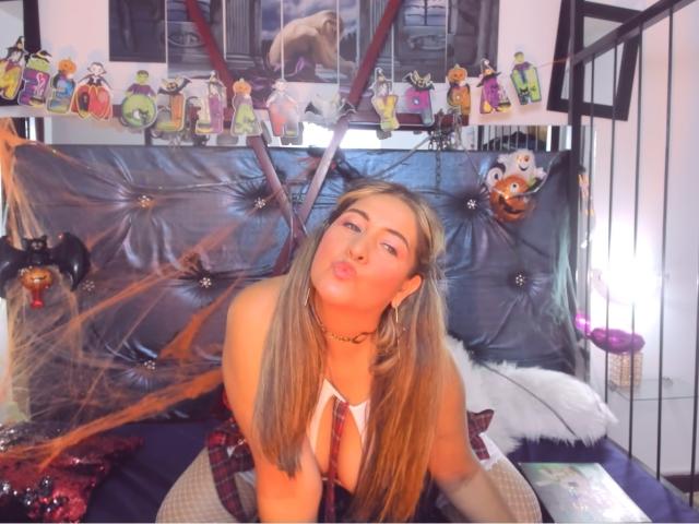 JessaOriz has long blonde hair, pouty lips, and is wearing a schoolgirl outfit with fishnet stockings, leaning on a black, tufted bed with her tits slightly visible.