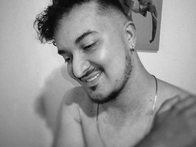 TheoOB has curly hair, a mustache, and a small nose ring. He's shirtless, smiling, and wearing a thin necklace. His ears are pierced, and he's looking down.