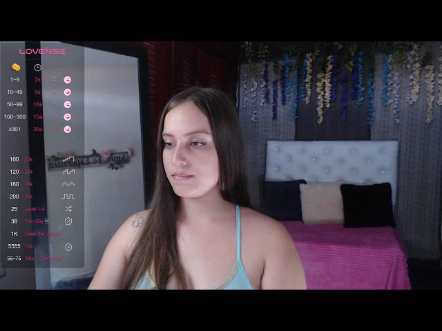 TaniaCooper69 with long brown hair, blue tank top, small tits, slight smile, tattoo on right shoulder, sitting in bedroom with pink bed, blue and white pillows.