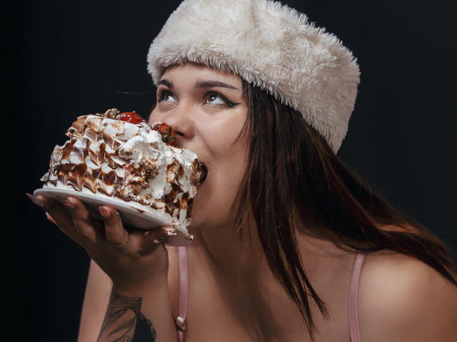 LallyyPop, com um chapéu branco fofo, comendo um bolo enorme coberto de creme, com a boca bem aberta, olhos castanhos olhando para cima, usando alças rosa, tatuagem na mão esquerda..