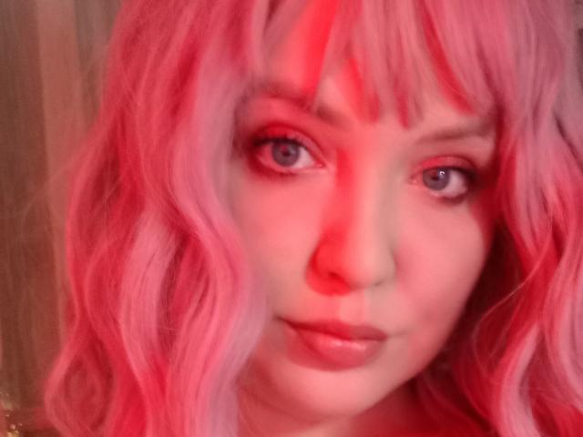AmeliaRey has pink wavy hair, red eyes, and red highlights on her face. She's looking at the camera with a slight smile, her pink lips visible.