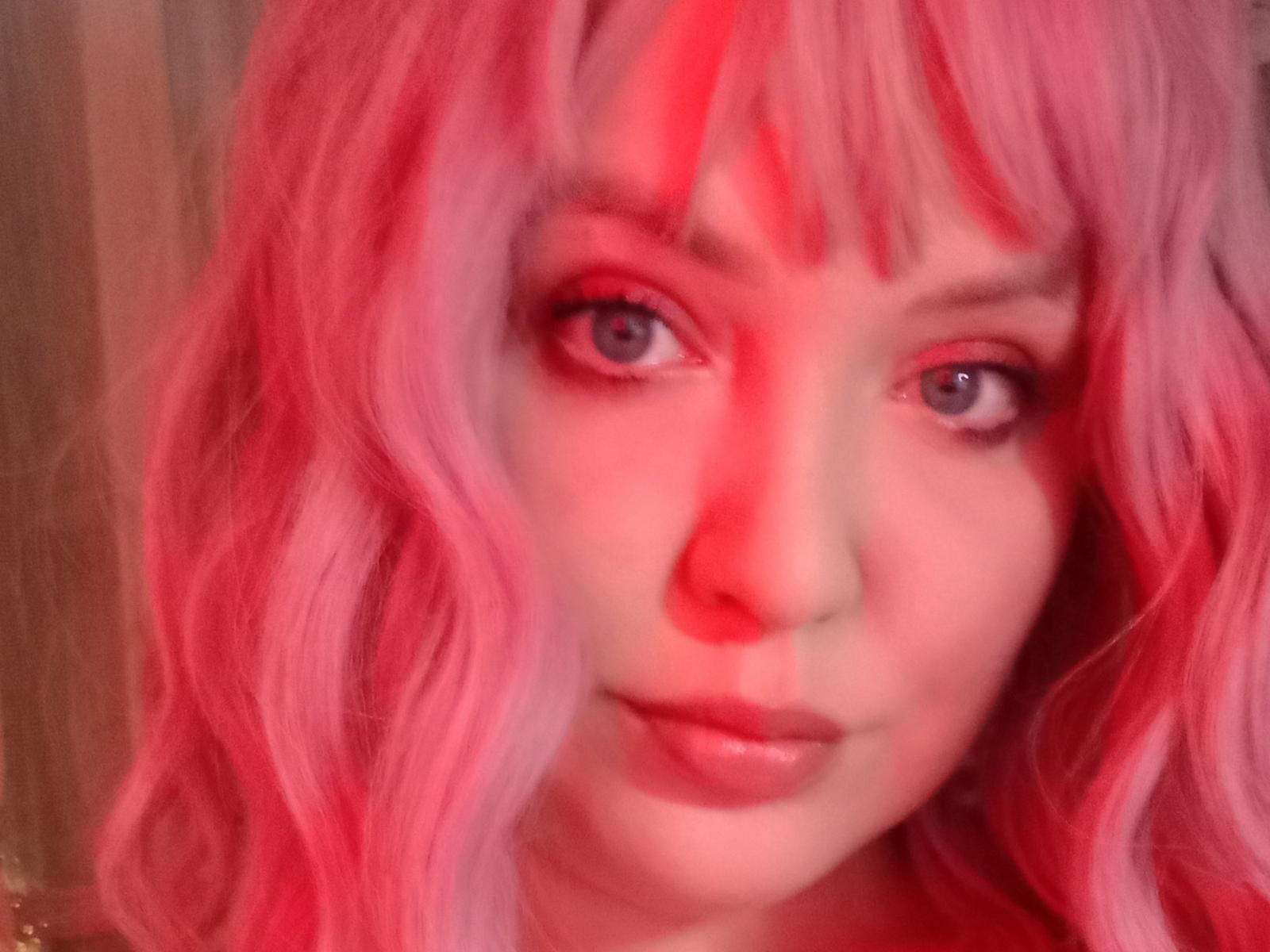 AmeliaRey has pink wavy hair, red eyes, and red highlights on her face. She's looking at the camera with a slight smile, her pink lips visible.
