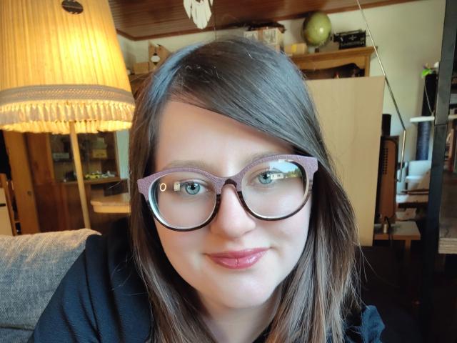 MelanieSweets-hot with purple glasses, pink lipstick, and straight brown hair, smiling slightly, wearing a black top, in a cozy room with a warm lamp.