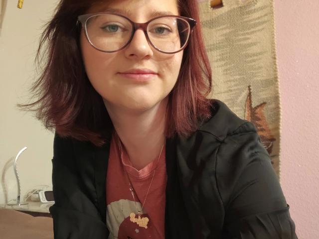 MelanieSweets-hot with red hair, glasses, black jacket, pink shirt, necklace, and slight smile, showing her tits subtly under her jacket, standing in a room with a pink wall.