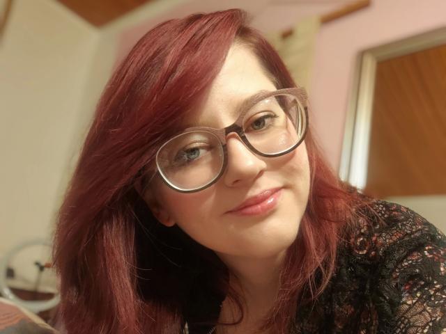 MelanieSweets-hot with red hair, wearing glasses, and a black lace top, smiling slightly with pink lips, looking cute and sexy.