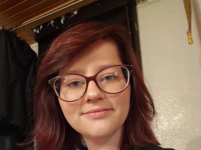 MelanieSweets-hot with red hair, glasses, and a slight smile, showing her face. She's got cute tits and a fair skin tone. She's wearing a black shirt.