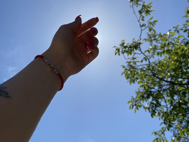 Olieris Hand mit rot lackierten Nägeln und einem roten Armband, die Sonne scheint im Hintergrund, sie streckt sich nach oben, grüne Blätter eines Baumes sind rechts zu sehen, klarer blauer Himmel, kleines Tattoo am Handgelenk..