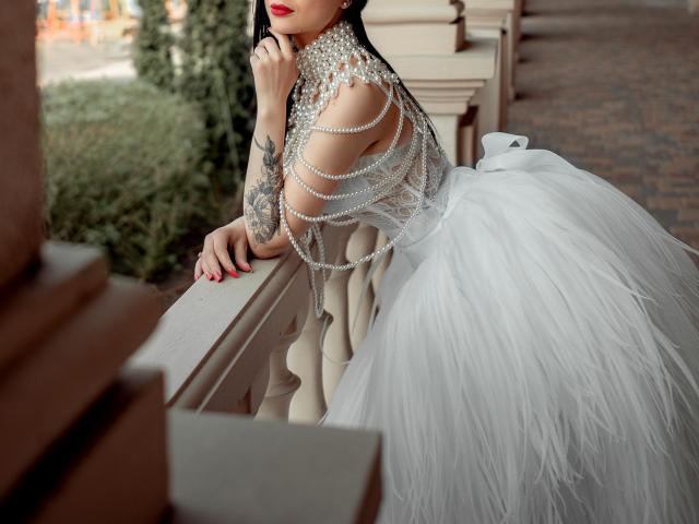 Olieri with tattoos, wearing a white dress with a massive, fluffy ass, covered in pearl chains, leaning on a beige railing, red lipstick, and manicured nails.