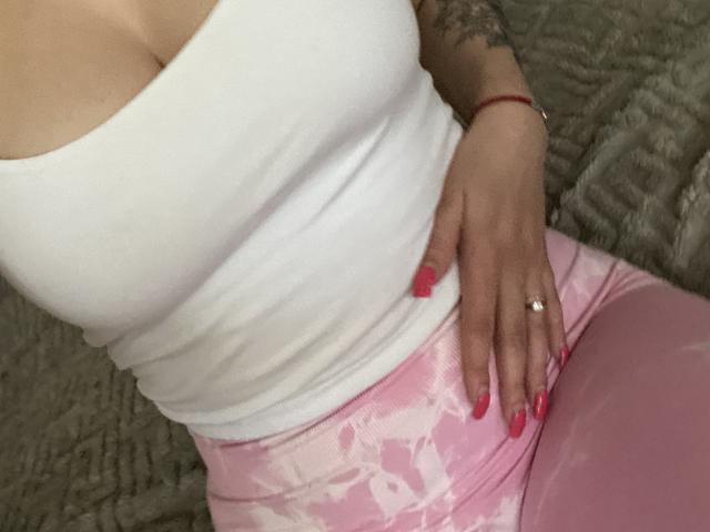 Olieri's tits in a tight white tank top, pink tie-dye leggings, red manicure, ring on her finger, tattoo on her arm, red bracelet, hand on her hip.