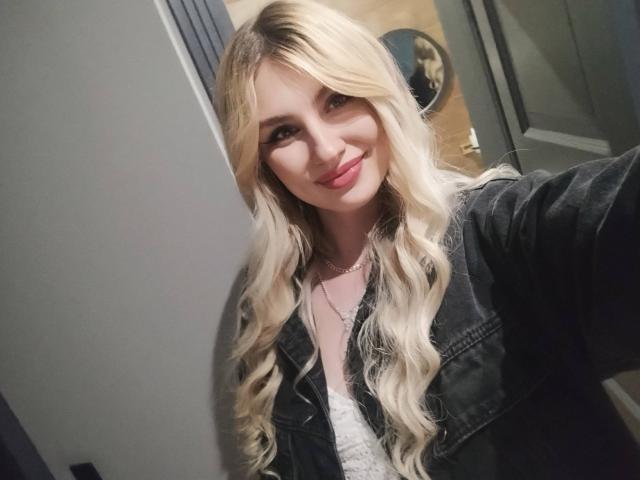 EvaLoona with long blonde wavy hair, smiling, wearing a black denim jacket over a white lace top, taking a selfie, showing her pink lipstick and cute face.