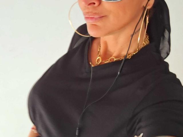 BriliantOne with big boobs, wearing a black sheer headscarf, gold hoop earrings, and a gold chain necklace, has a slightly pouty face and is holding a phone.