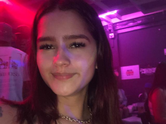 MelodyMansson has brown hair, full lips, and a subtle smile, with long eyelashes and a gold necklace, under pink and red lighting. Her face is centered, slightly blurred.