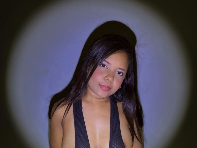 MelikaSweet has dark straight hair, light brown skin, and small tits in a low-cut black top. She's looking at the camera with a slight smile.