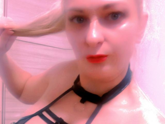 Blonde MonicaMiss with shiny skin, red lips, black strappy top, holding hair, blurry pink-tinted selfie, visible breasts, black choker, looking at camera with slightly open mouth.