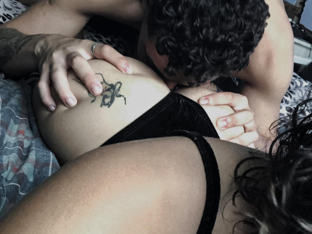 CasalMisterioso's tattooed arm rests on their black lace panties while another person with curly hair, nose buried in their pussy, grips their ass. Tattoo: cross with arrow.