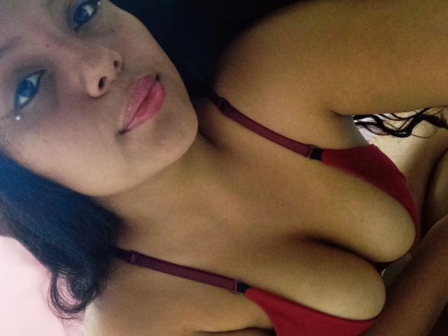 KonyBella with pouty pink lips, wearing a red bra, showing her big boobs and cleavage, has dark hair and a small beauty mark on her left cheek.