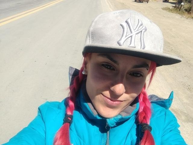 Amy, mit roten Zöpfen, blauer Jacke und einer grauen New York Yankees Mütze, lächelt in die Kamera, im Hintergrund ist eine Straße zu sehen. Ihre Brüste sind nicht sichtbar..