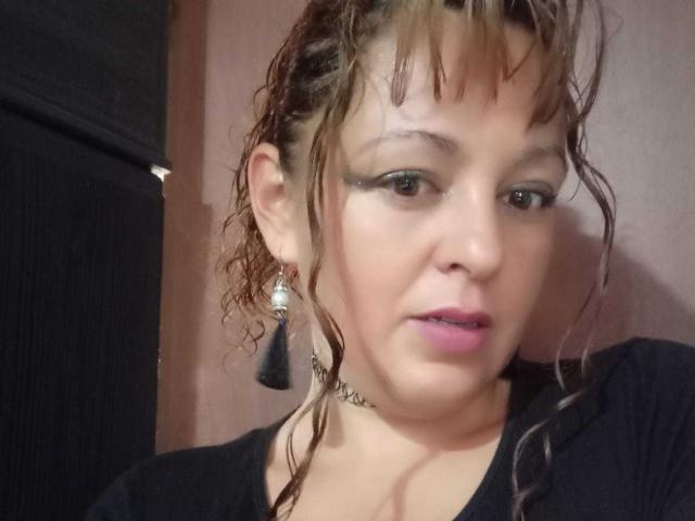 AmandaHotL has curly brown hair, black eyeliner, pink lipstick, black t-shirt, and black tassel earrings, with a choker necklace, looking slightly open-mouthed in a selfie.
