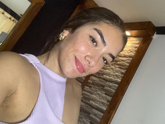 SharonKlart with wet brown hair, pink glossy lips, white ribbed top, gold earrings, and a subtle smile, showing her shiny face and small, perky tits.
