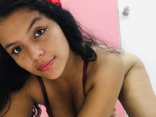 KonyBella with dark curly hair, red lipstick, and a nose piercing, wearing a red bra, smiling, showing her tits, and a slight view of her ass.