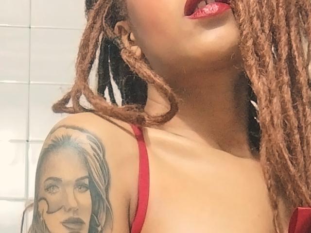 KendallQuinn with long brown braids, red satin lingerie, bow, and gloves, showing tits and tattoos, standing in a bathroom, looking up with red lipstick.