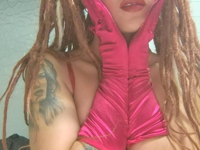 KendallQuinn with pink, shiny, satin gloves, red lipstick, and brown dreadlocks, showing a tattoo on her right arm, arms raised, showing her tits.