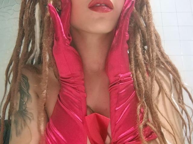 KendallQuinn with pink satin gloves, red lipstick, and brown dreadlocks, showing a tattoo on her left arm, her tits slightly visible through the cleavage.