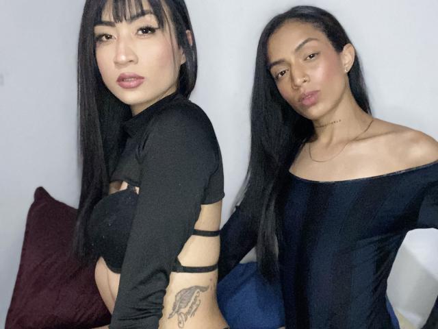 AfrickXDakotta with long black hair, wearing a black crop top, showing a tattoo on her arm, and another AfrickXDakotta in a black off-shoulder top, both looking at camera.