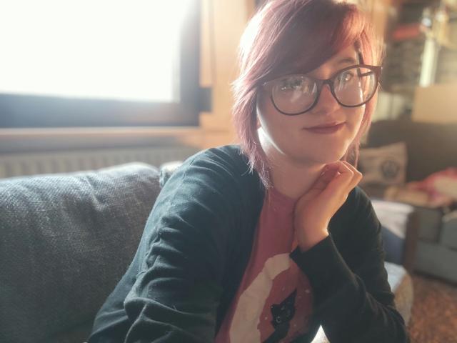 MelanieSweets-hot with red hair, glasses, black cardigan, pink shirt, and hand on chin, smiling softly, sunlight on face, cozy indoor setting.