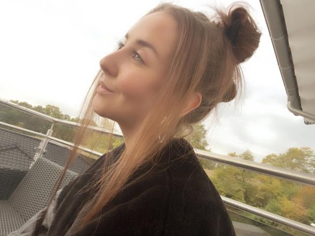 Kia-rey-hot with her brown bun and long hair, smiling slightly, wearing a black shirt, standing on a metal balcony with a cloudy sky and green trees in the background.