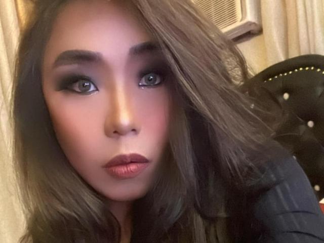 MissSultryAya's face, featuring big blue eyes, full pink lips, and long brown wavy hair, with a serious expression, wearing a black top, in a slightly blurry selfie.