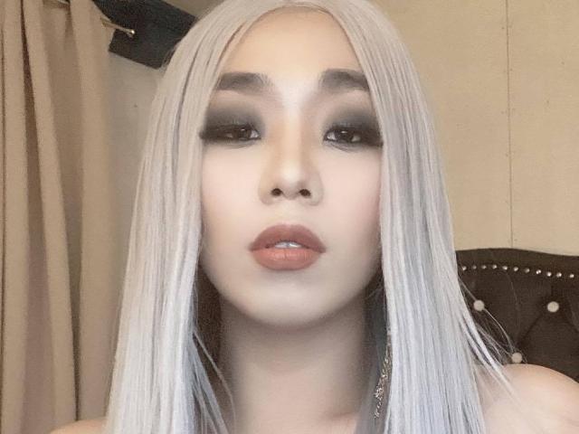 MissSultryAya heeft bleke huid, lang, steil, platina blond haar, donkere, smoky oogmake-up en volle, roze-bruine lippen. Ze kijkt recht in de camera met een neutrale uitdrukking..