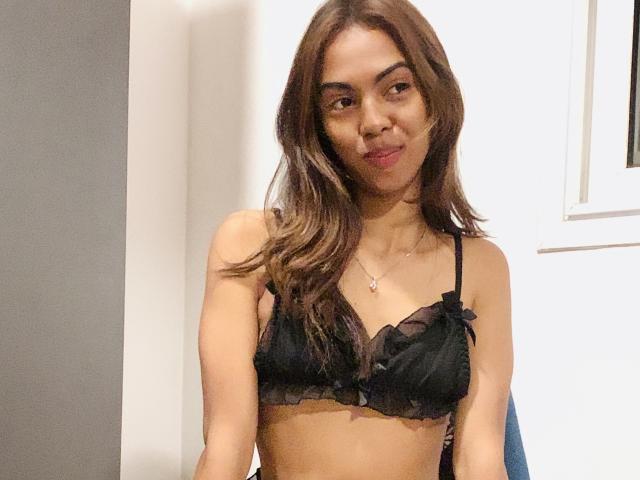 CarlaFinger, with brown wavy hair, wearing a black bra, has small tits, a cute face, and a subtle smile, standing indoors. She's wearing a silver necklace.