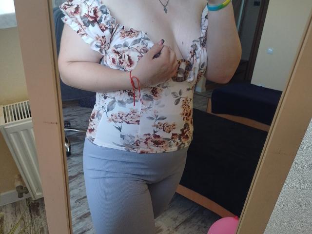 Leiliia's floral top with ruffled shoulder, exposing her boobs, wearing gray leggings, red bracelet, and colorful wristband, reflected in a mirror. Her hand covers her nipple.