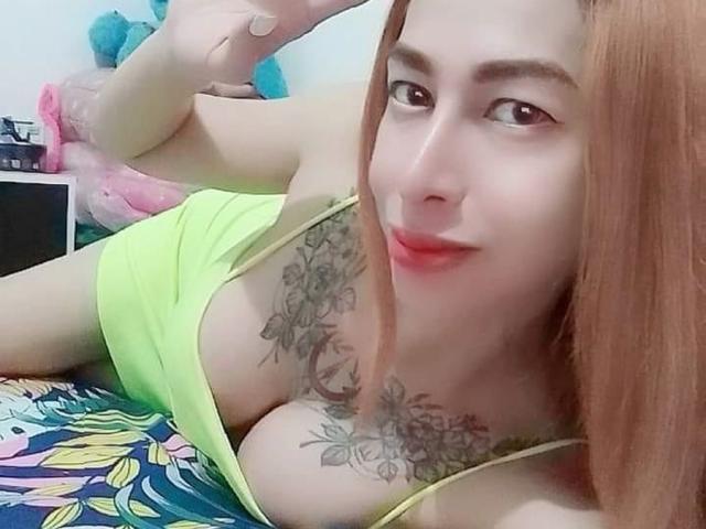 Eine verführerische, attraktive Frau mit roten Haaren, großen Brüsten und Tattoos, liegt auf einem bunten Bett, trägt ein neon-gelbes Oberteil, lächelt und zeigt ein Friedenszeichen..