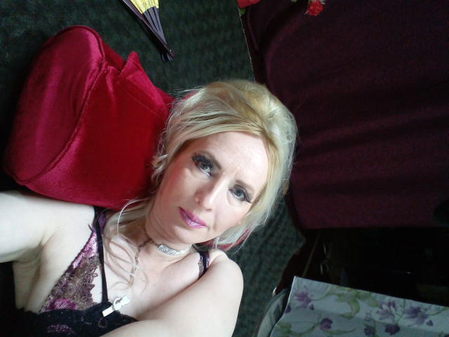 Blonde EroLayer with purple lipstick, black lace lingerie, and a silver choker, lying on a dark carpet, red bag beside her, purple umbrella in the background.