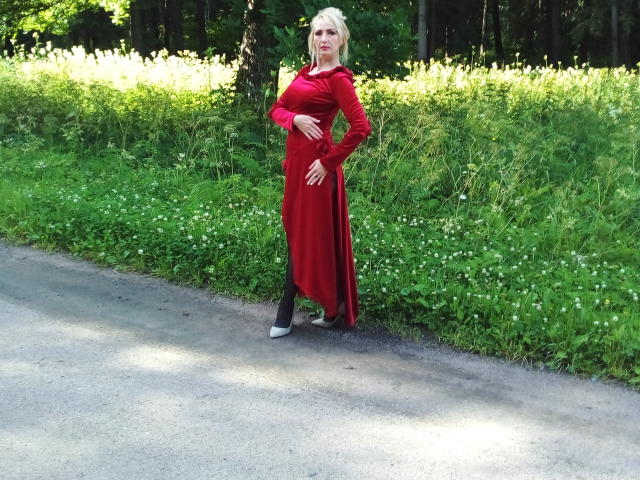 EroLayer stands on a gravel path, wearing a red dress, black pants, and white shoes, with blonde hair, holding her hand to her belly, showing her ass.